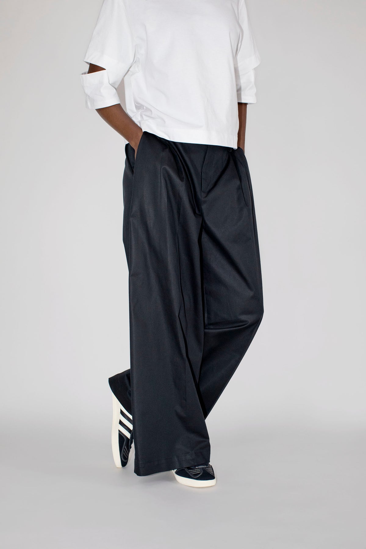 A model from the waist down wearing loose, wide-leg black trousers, a white t-shirt, and black sneakers with white stripes, standing against a light gray studio background.