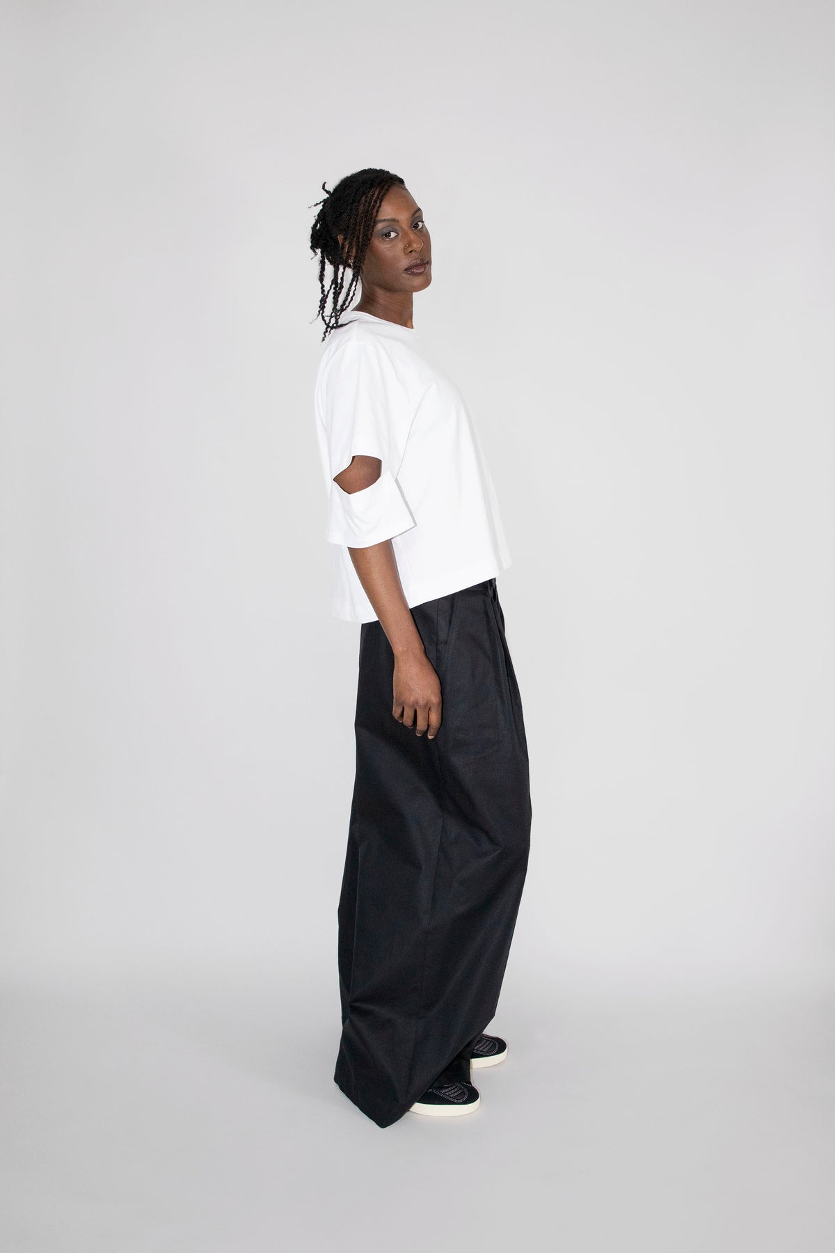 A model stands in profile wearing a white short-sleeve top with a cutout detail on the sleeve and black wide-leg trousers, set against a plain light gray studio background.