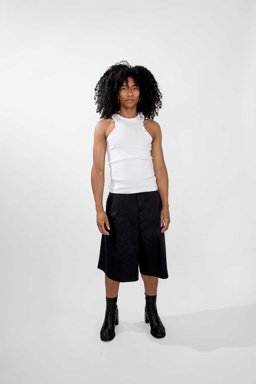 A model with curly black hair stands against a light gray studio background wearing a white sleeveless mock-neck top, black wide-leg shorts, and black ankle boots.