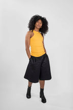 A model with curly black hair stands against a light gray studio background wearing a bright yellow sleeveless ribbed tank top, black wide-leg shorts, and black ankle boots.